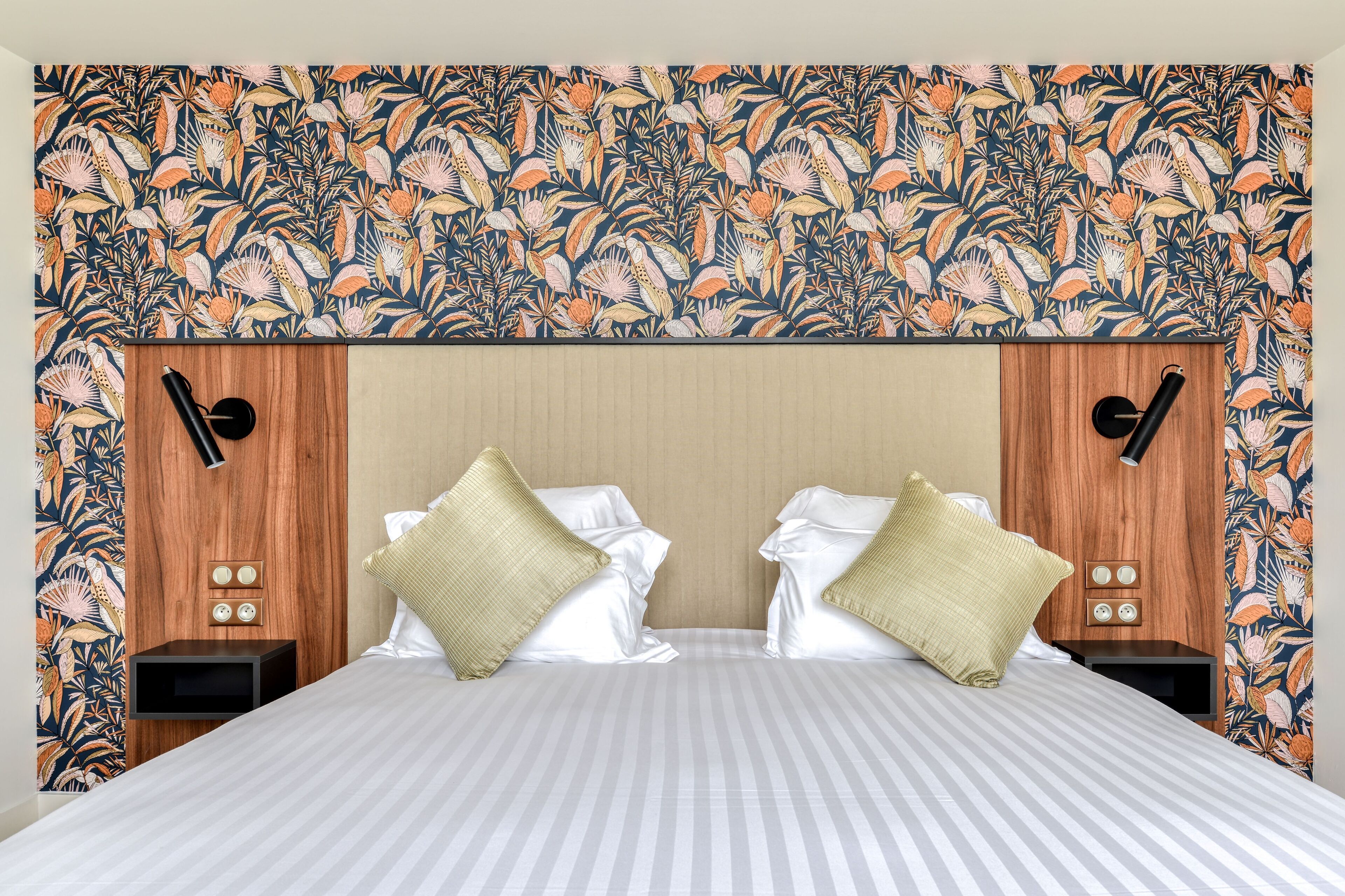Une chambre d’hôtel avec un papier peint à motifs floraux, une couvre-lit à rayures, deux tables de chevet avec des lampes et deux coussins décoratifs.