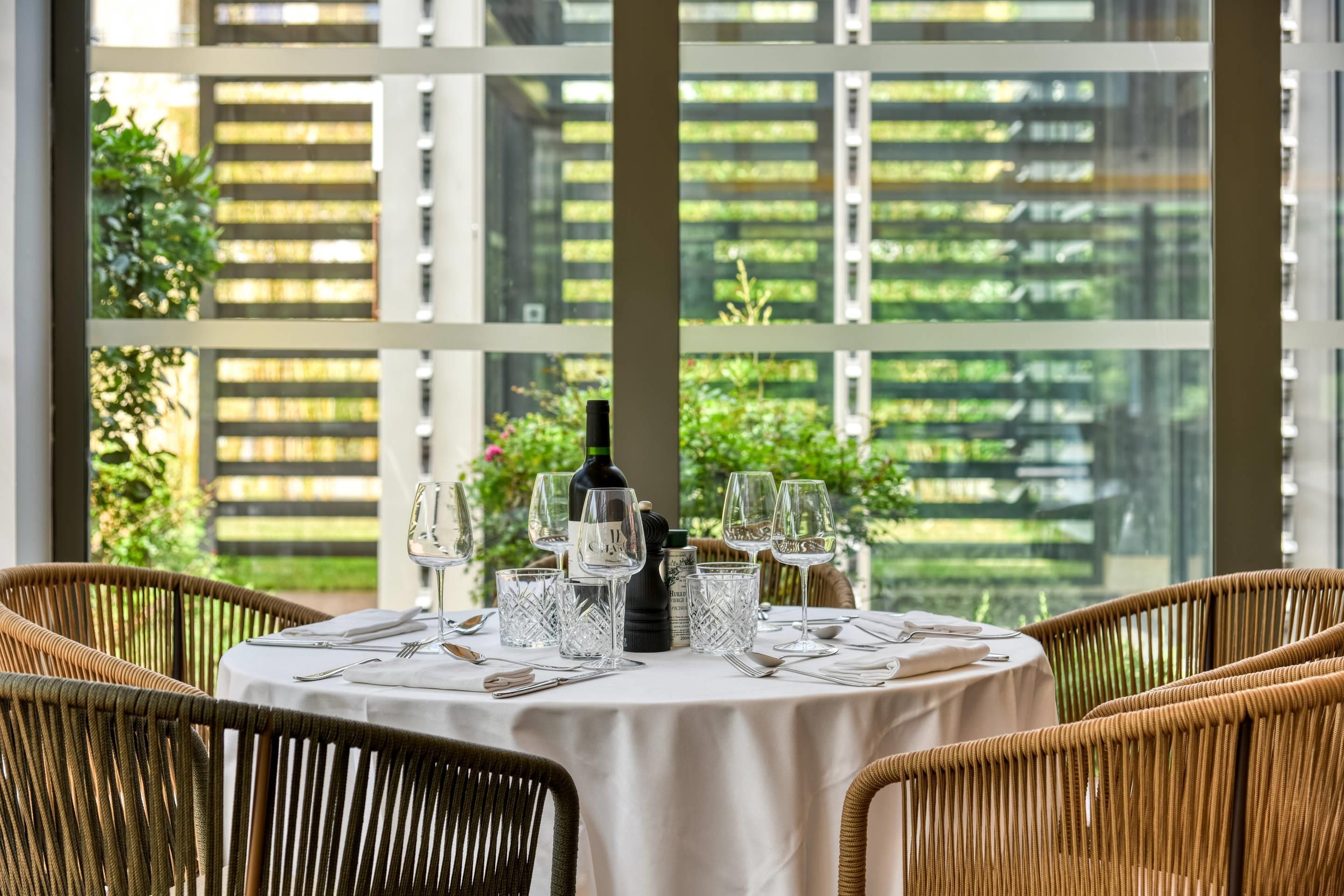 Une table de salle à manger ronde, recouverte d’une nappe blanche, entourée de chaises en osier, mise avec des verres à vin, une bouteille et des couverts.