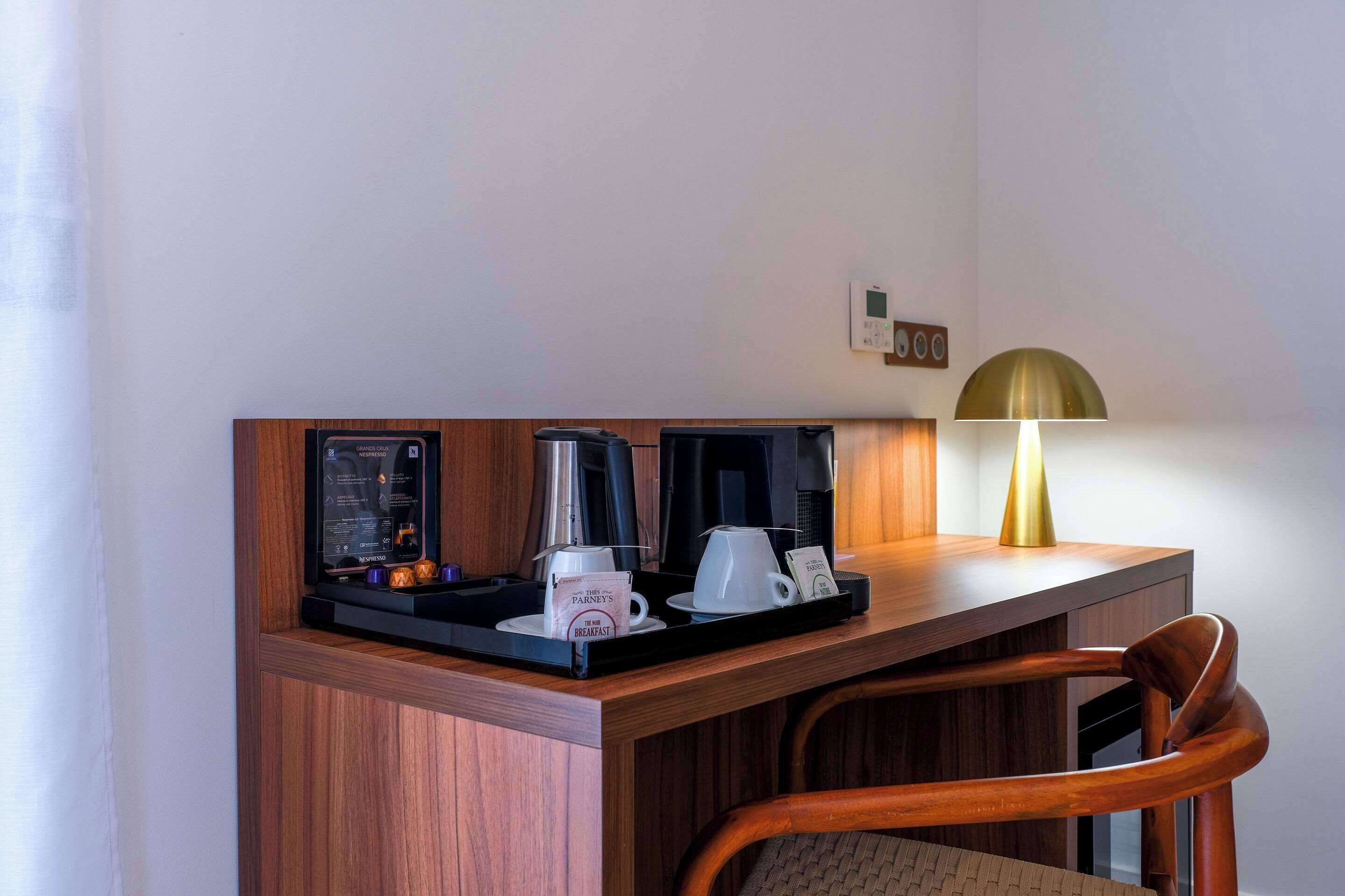 Une chambre d’hôtel avec un bureau en bois, une machine à café, une tasse de café, une lampe et un menu encadré.