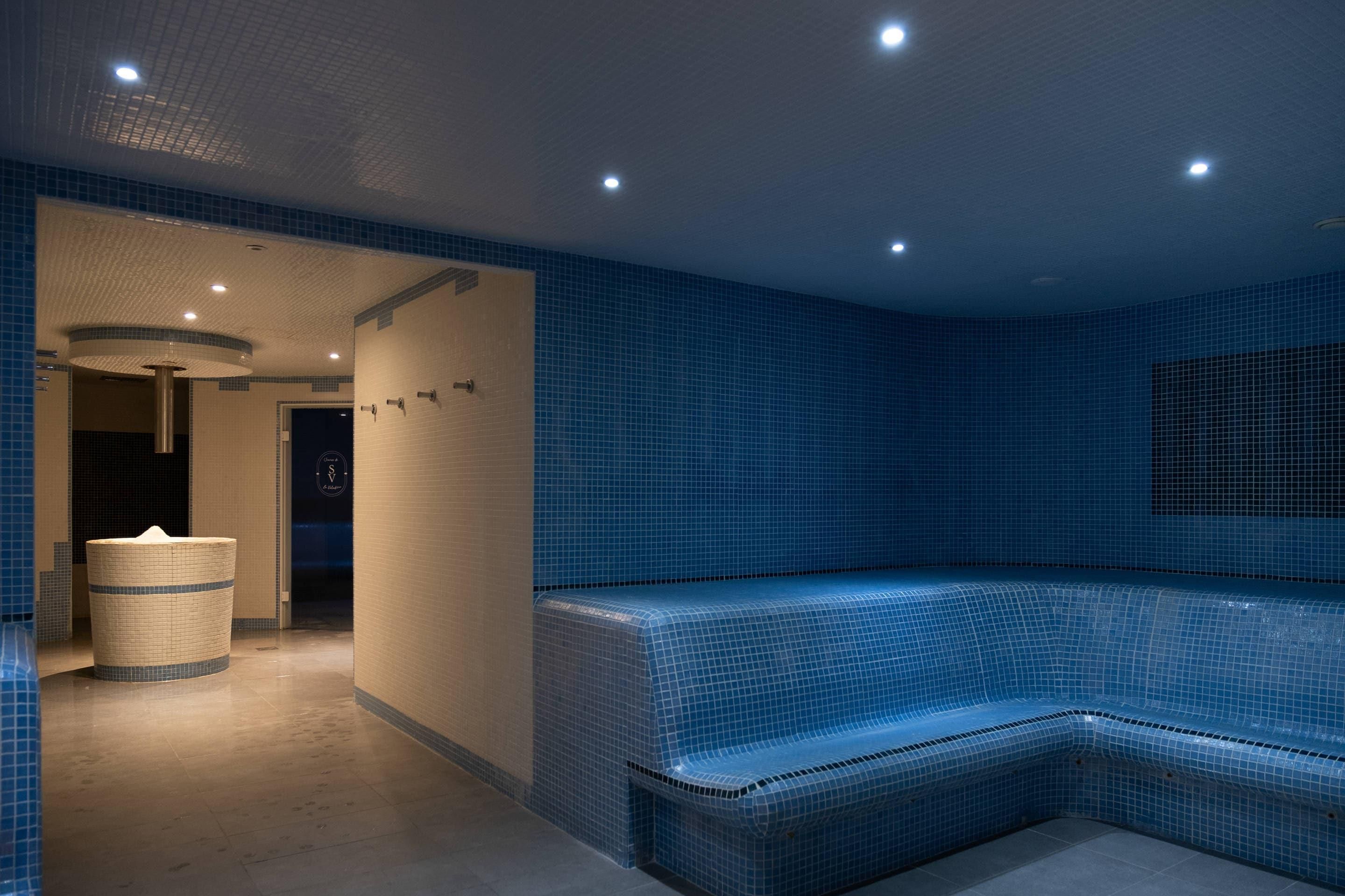 Uma área de spa com azulejos azuis, espaço para sentar e um grande recipiente cilíndrico.
