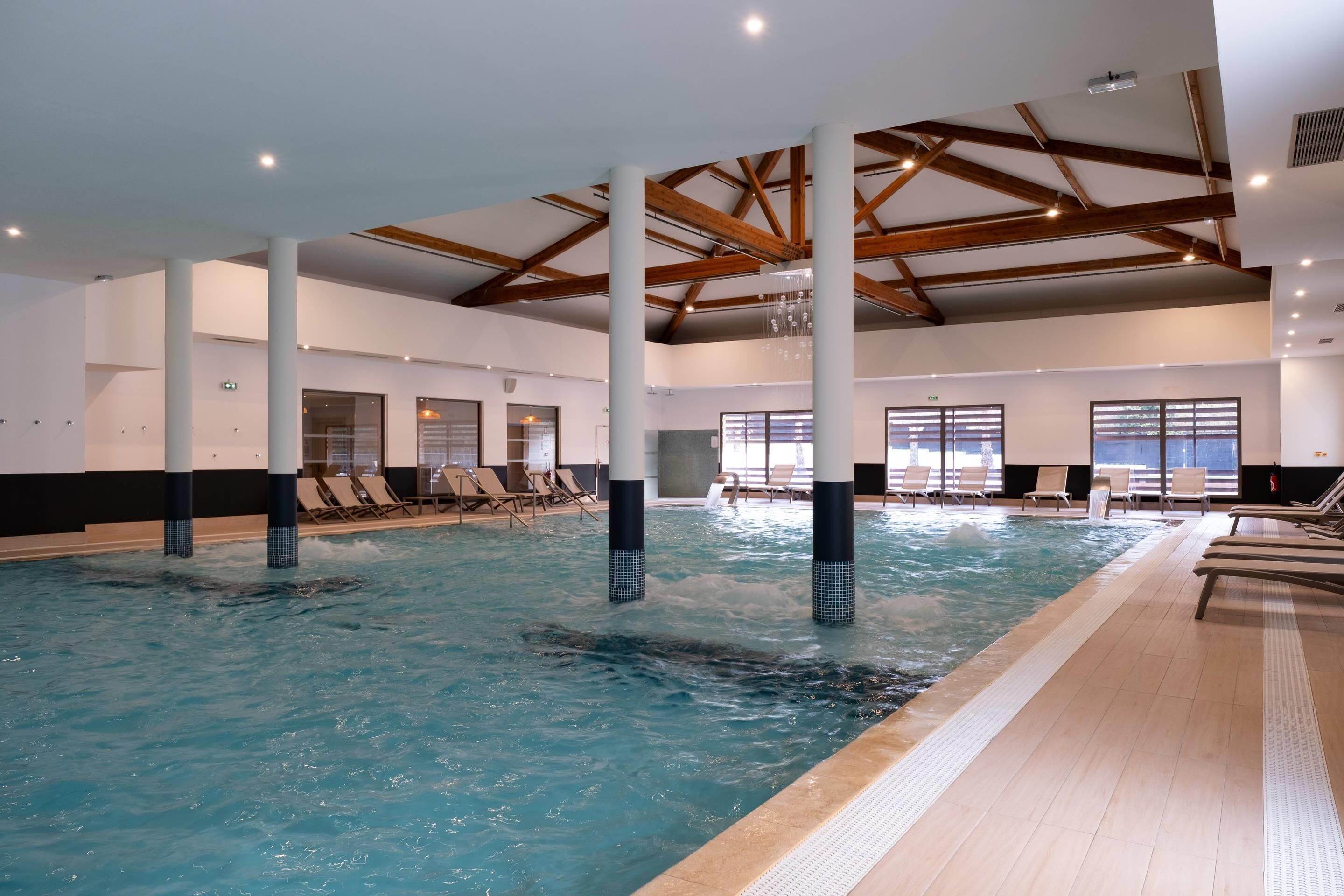 Une piscine intérieure à l’eau claire, avec des poutres en bois au plafond et des chaises longues disposées le long du bord de la piscine.