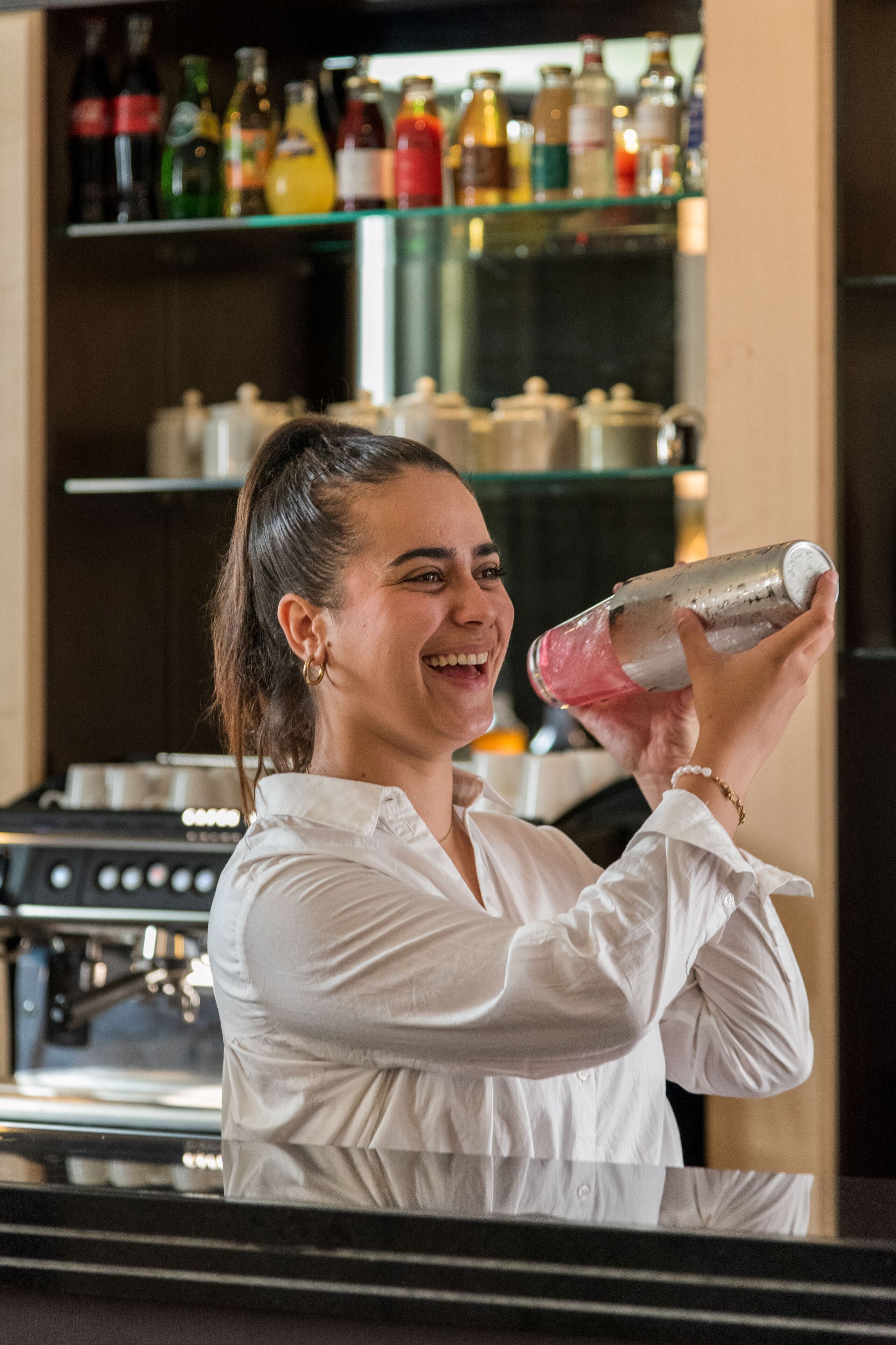 A person holding a cocktail shaker in a kitchen setting with shelves of bottles and kitchen appliances in the background.