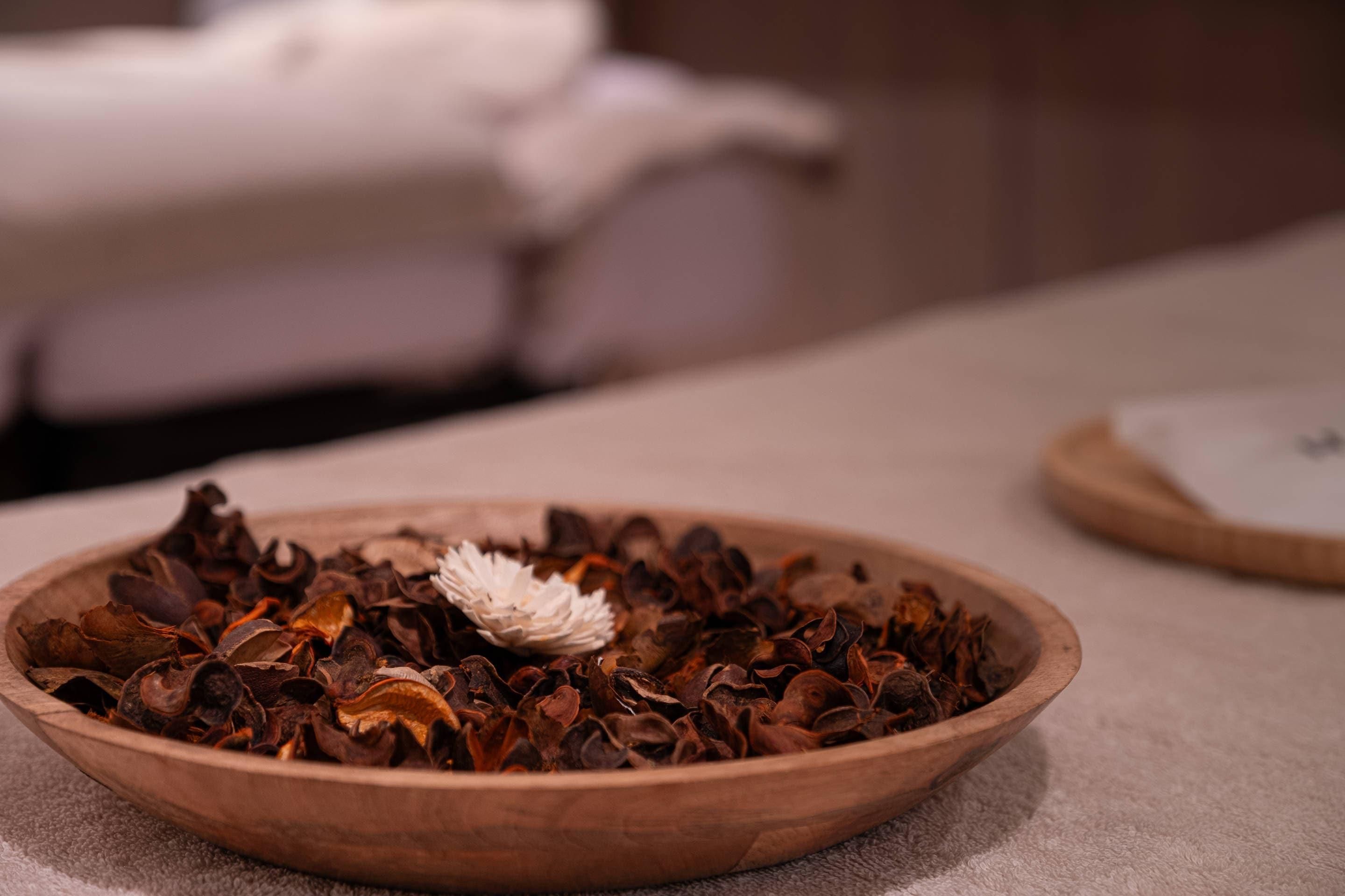 A wooden bowl filled with dried leaves and a white flower, placed on a table.