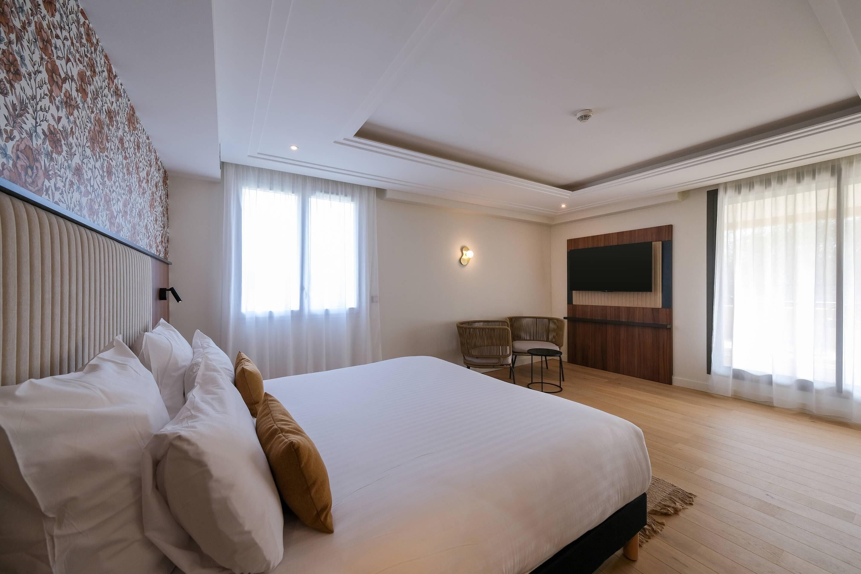 Quarto de hotel com uma cama grande, televisão, área de estar com cadeiras e janela com cortinas.