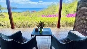 Una veranda coperta con due sedie in vimini, un tavolo con un vaso e una ciotola, e vista sull'oceano e sul verde.