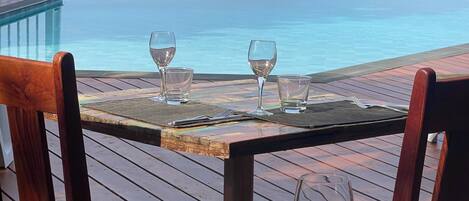 Um deck de madeira com mesa e cadeiras, dando para uma piscina e o mar.