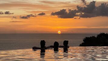 Un coucher de soleil sur l’océan, avec les silhouettes de personnes se détendant dans une piscine à débordement.