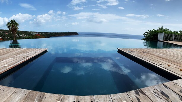 Una piscina a sfioro blu brillante, con bordi in legno, immersa nel verde e con vista sull'oceano.
