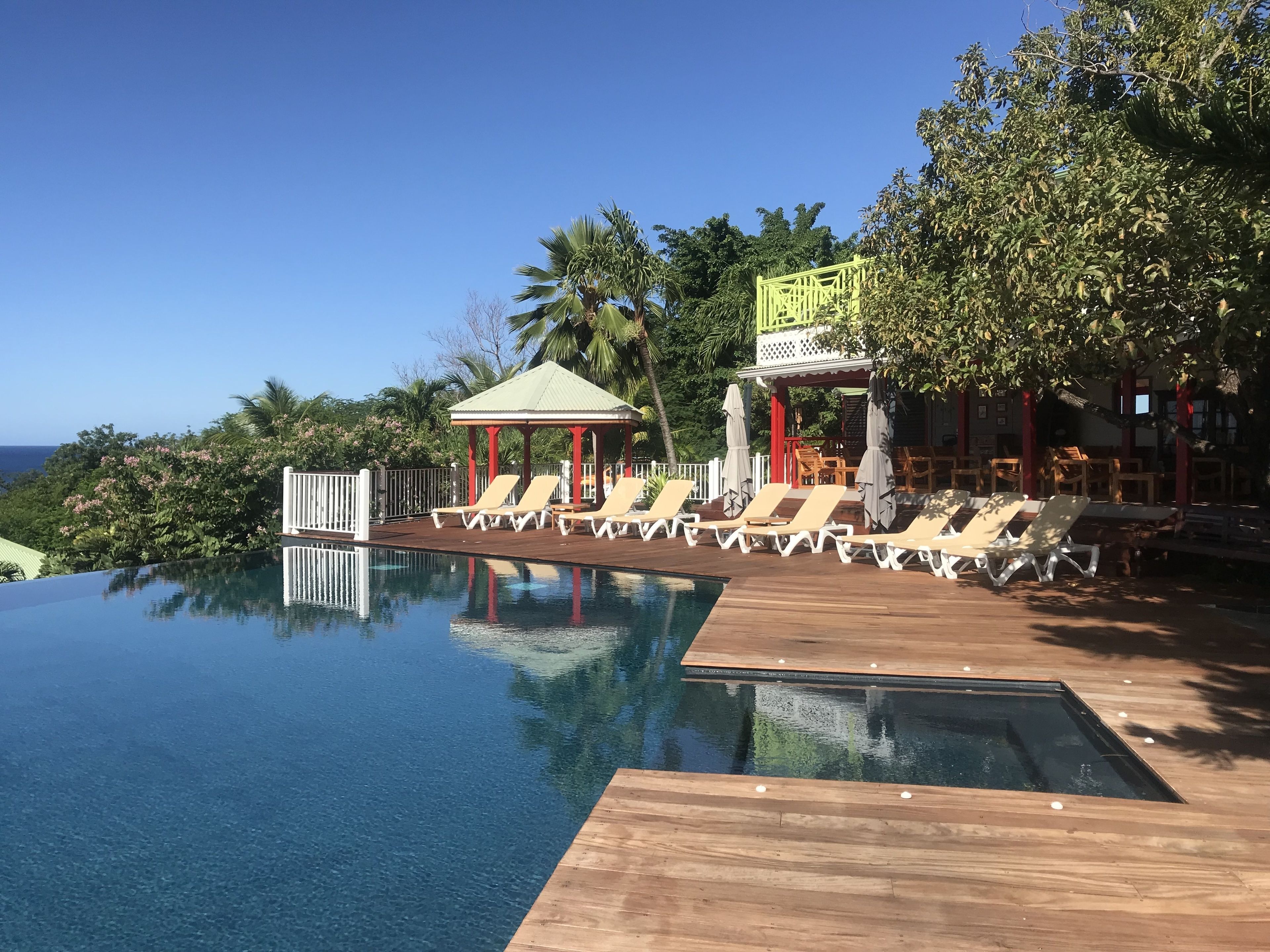 Una piscina infinita azul cristalina con cubierta de madera y sillas de descanso, rodeada de exuberante vegetación y vistas al océano.