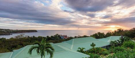 A coastal property with multiple green-roofed buildings, lush greenery, and a view of the sea at sunset.