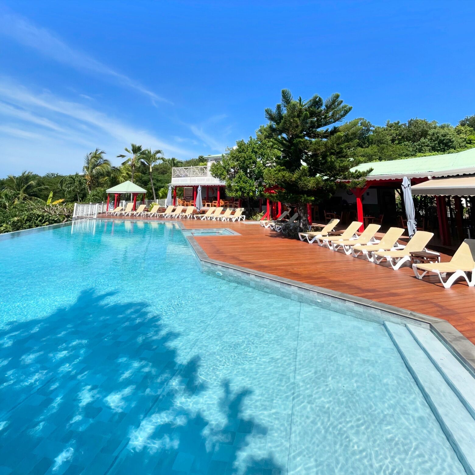 A clear blue pool with lounge chairs and umbrellas, surrounded by tropical vegetation and a wooden deck.