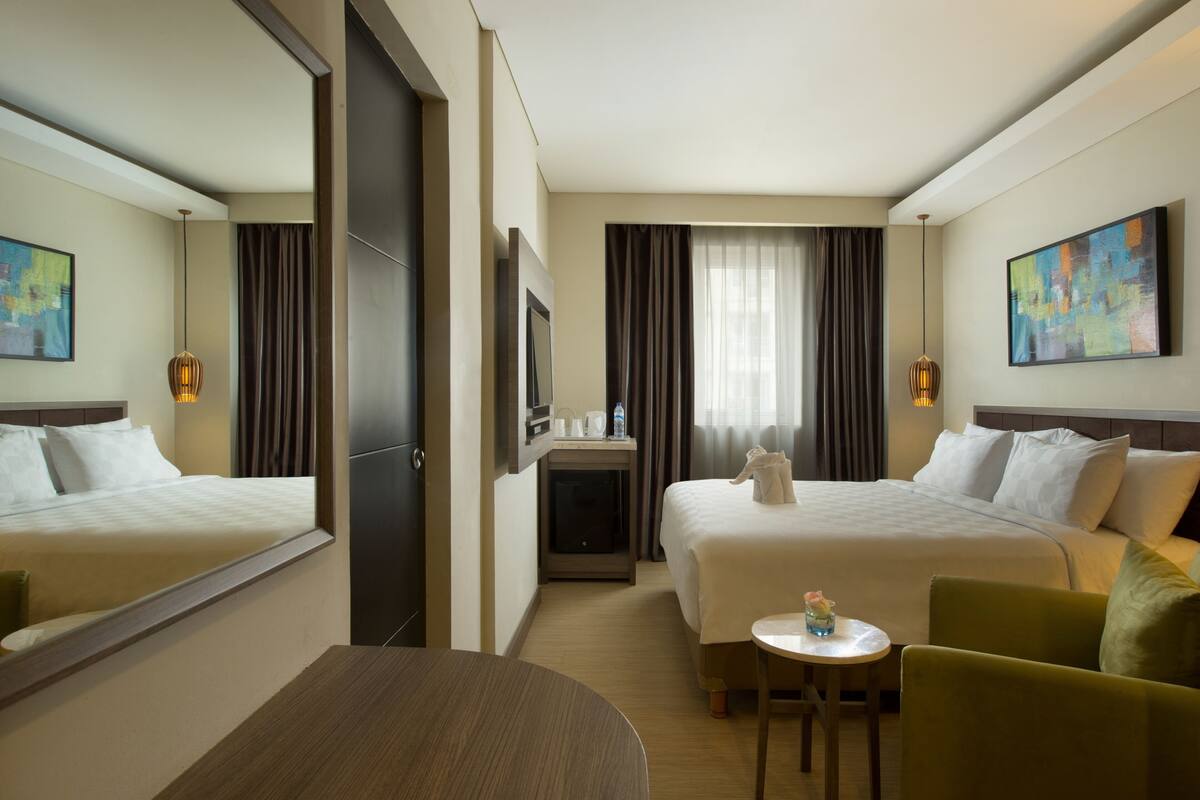 A hotel room with two beds, a small table, a chair, a desk, a mirror, and artwork on the walls.