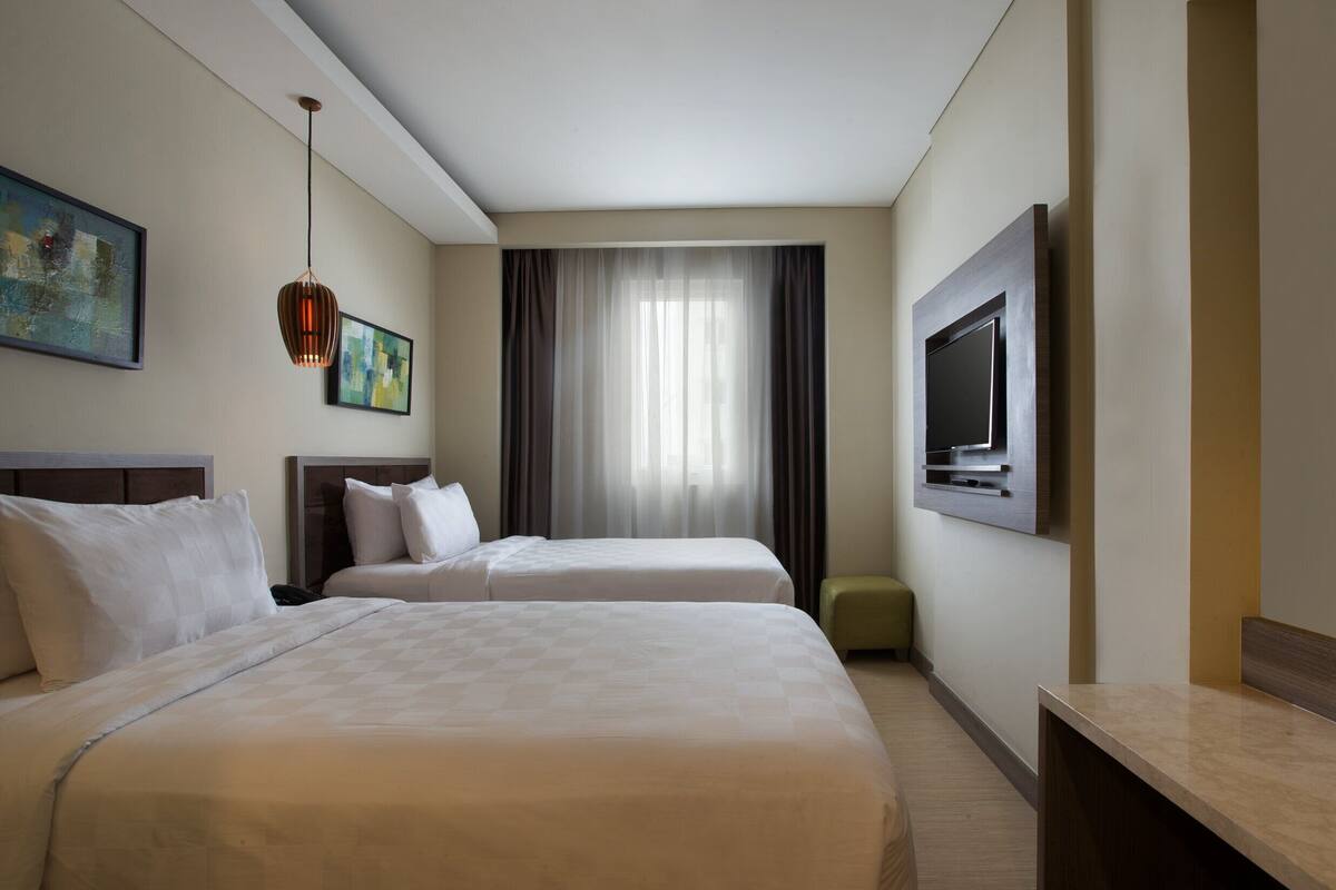 A hotel room with two beds, a flat-screen TV, a painting, and a window with curtains.