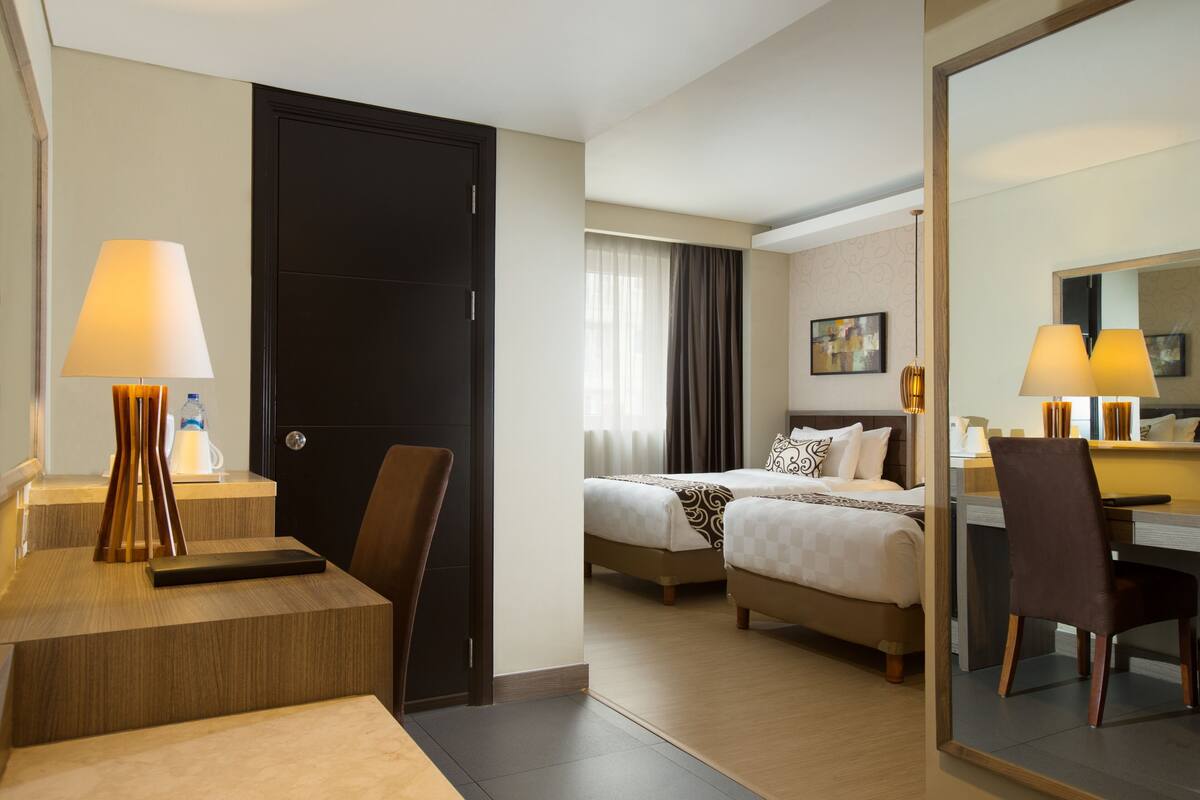 A hotel room with a desk, two beds, a chair, a mirror, and a door.