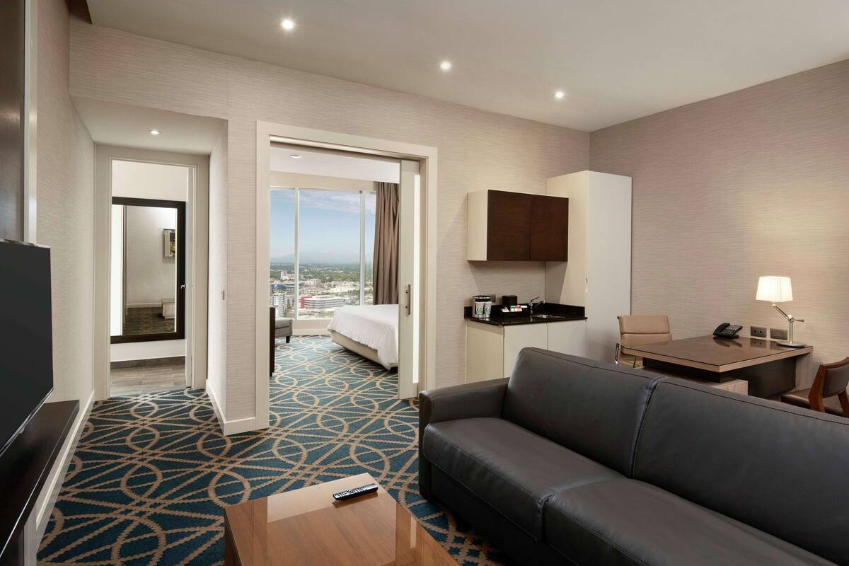 Ein Hotelzimmer mit einem großen Bett, einer Couch, einem Schreibtisch und einem Fernseher. Das Fenster bietet einen Blick auf die Stadt.
