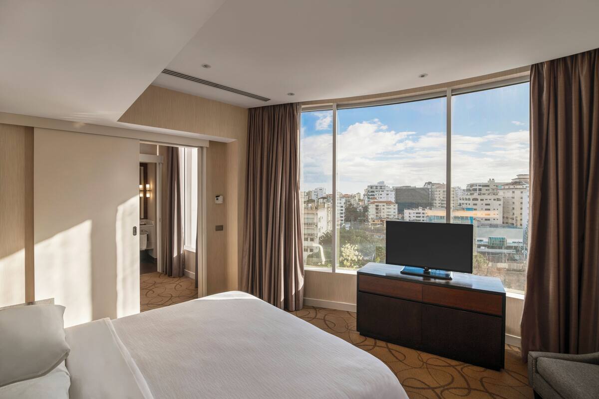 Ein Hotelzimmer mit einem großen Bett, einem Fernseher auf einer Holzkommode und Blick auf die Stadt durch große Fenster.