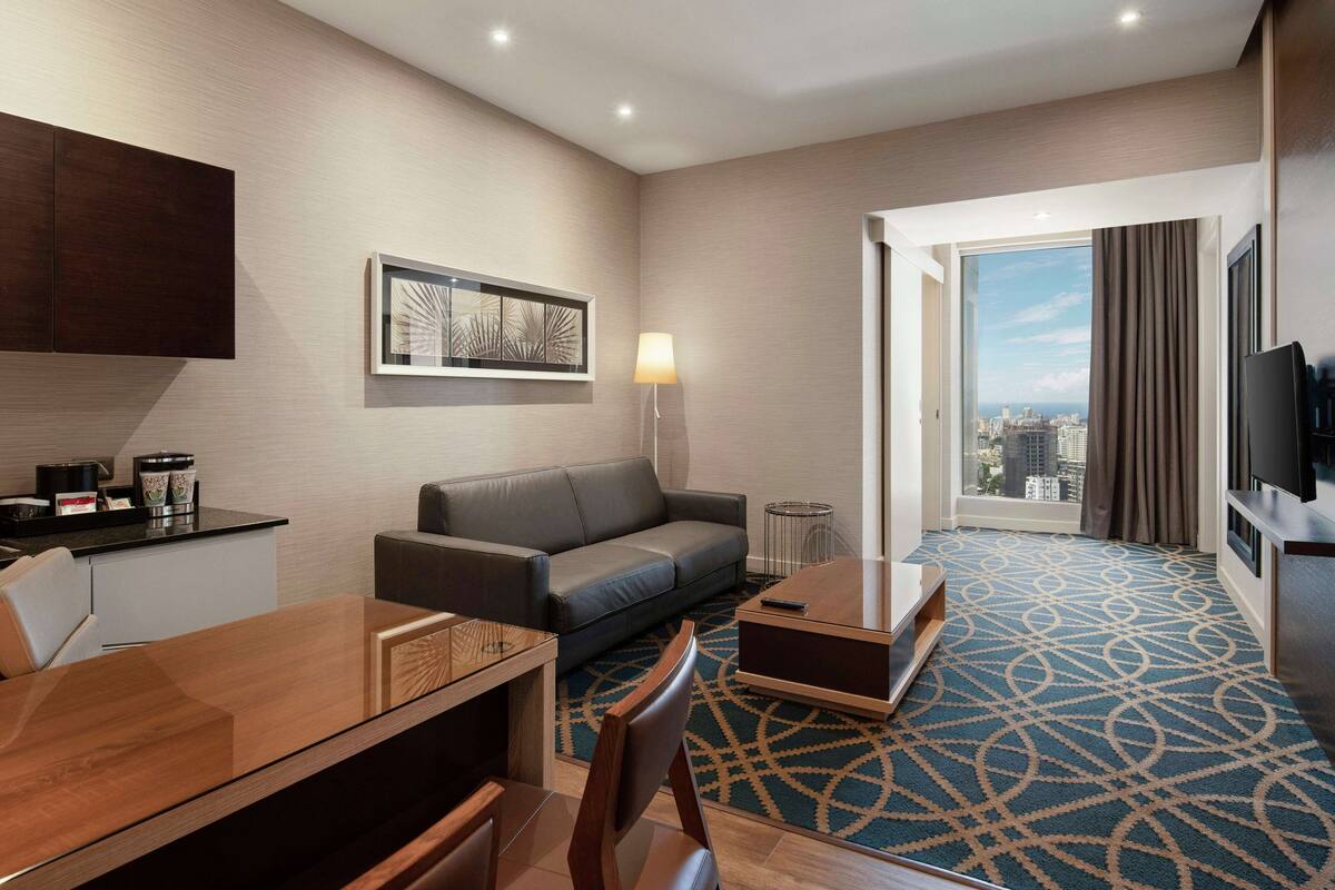 Ein modernes Hotelzimmer mit einer Couch, einem Esstisch, einem Fernseher, Blick auf die Stadt durch das Fenster und einem gemusterten Teppich.