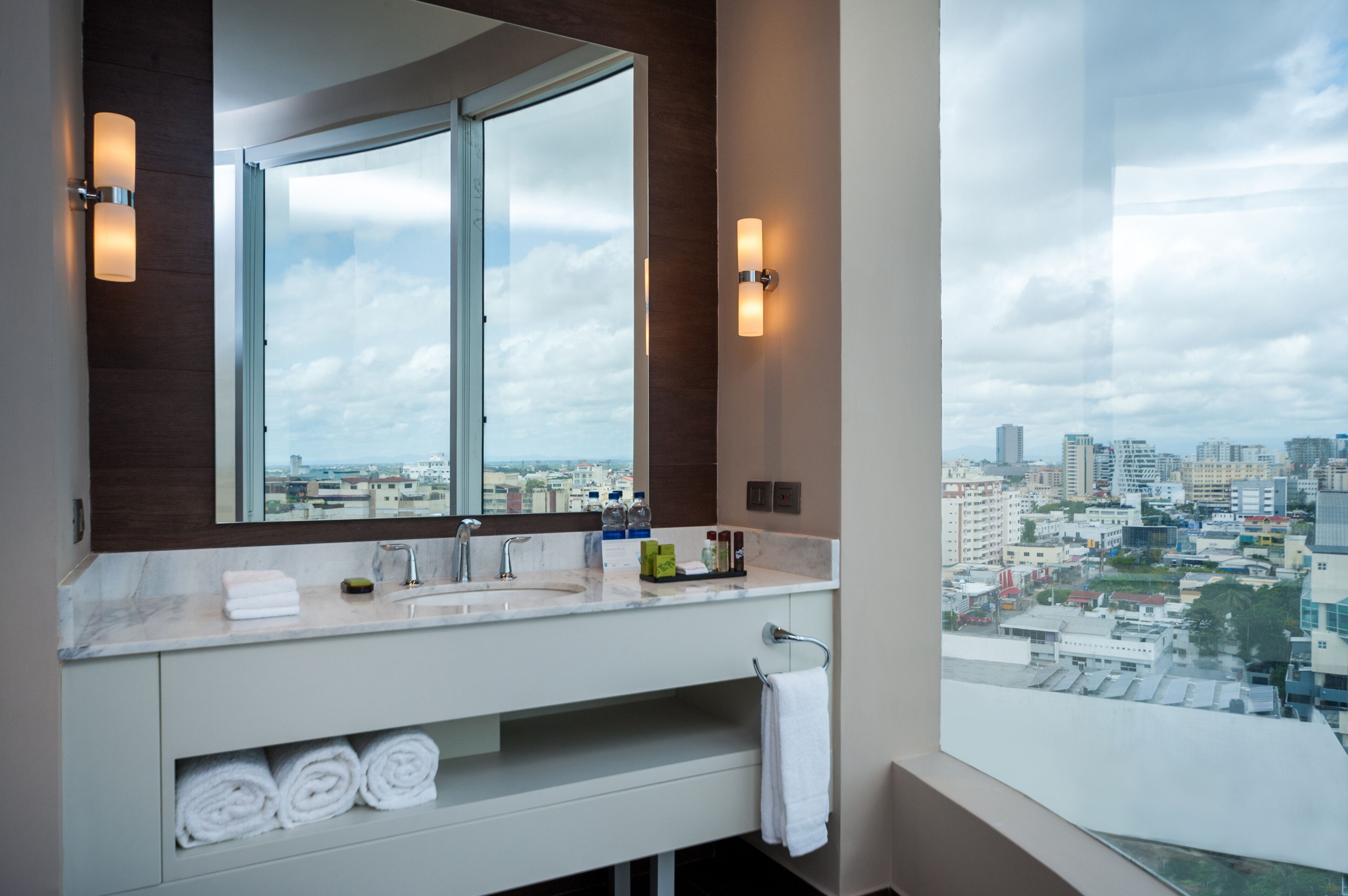 Une salle de bain moderne avec un grand miroir, deux vasques et une vue sur le paysage urbain, visible à travers une grande fenêtre.