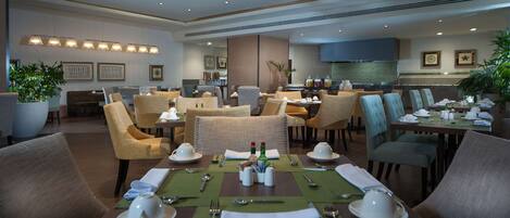 A hotel dining area with multiple tables set for dining, chairs, and a kitchen in the background.