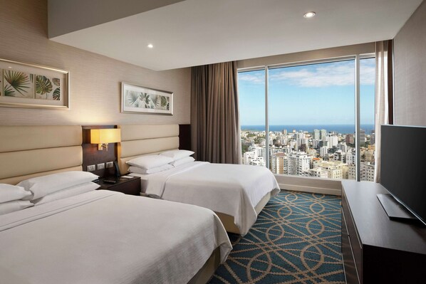 Habitación de hotel con dos camas, un televisor, un ventanal amplio con vistas a la ciudad y obras de arte en las paredes.