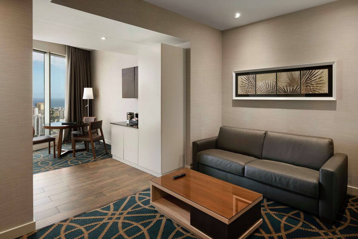 Ein modernes Hotelzimmer mit einer Ledersofa, einem Couchtisch, einem Essbereich mit Tisch und Stühlen sowie einem großen Fenster mit Blick auf die Stadt.