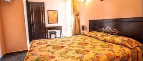 A hotel room with a floral bedspread, a dark wooden headboard, a small desk, and a framed picture on the wall.