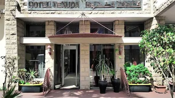 Entrada de hotel con un arco central, plantas en macetas a cada lado y un letrero sobre la entrada.