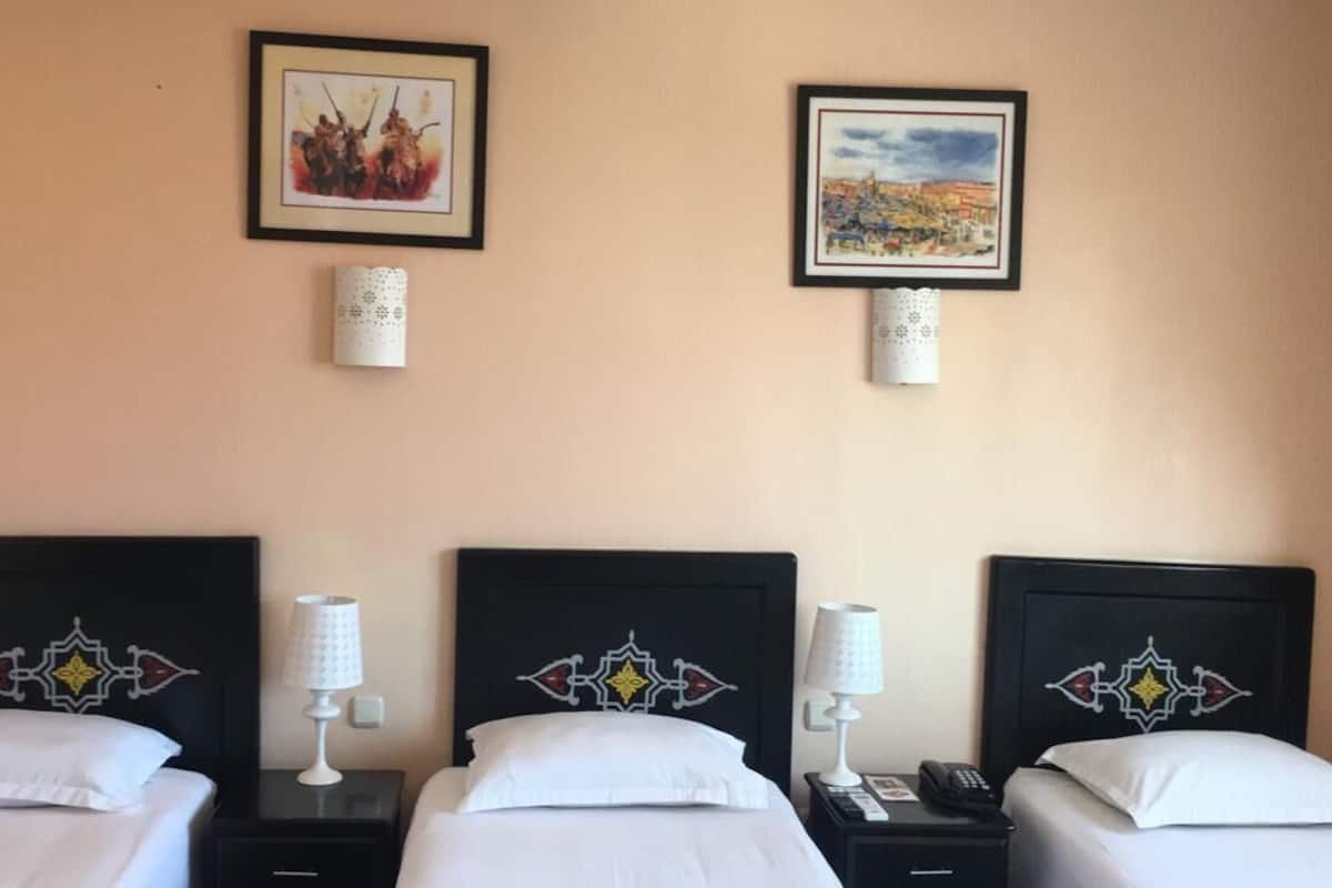 A hotel room with three beds, each with a black headboard featuring a geometric design. There are two wall-mounted lamps on either side of the beds.