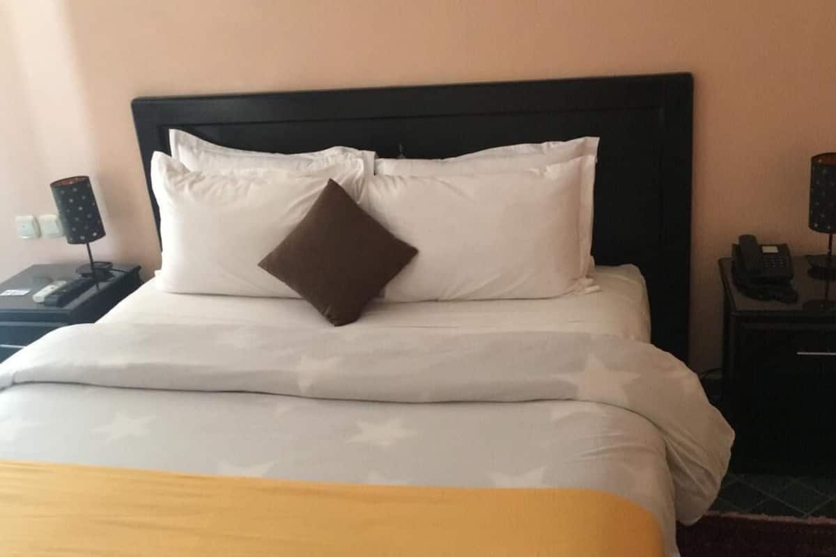 A hotel room with a neatly made bed, a brown accent pillow, two bedside tables with lamps, and a nightstand with a phone.