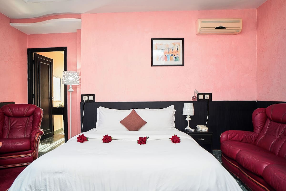 A hotel room with a bed, two red leather chairs, a nightstand with a lamp, and a wall-mounted air conditioner.