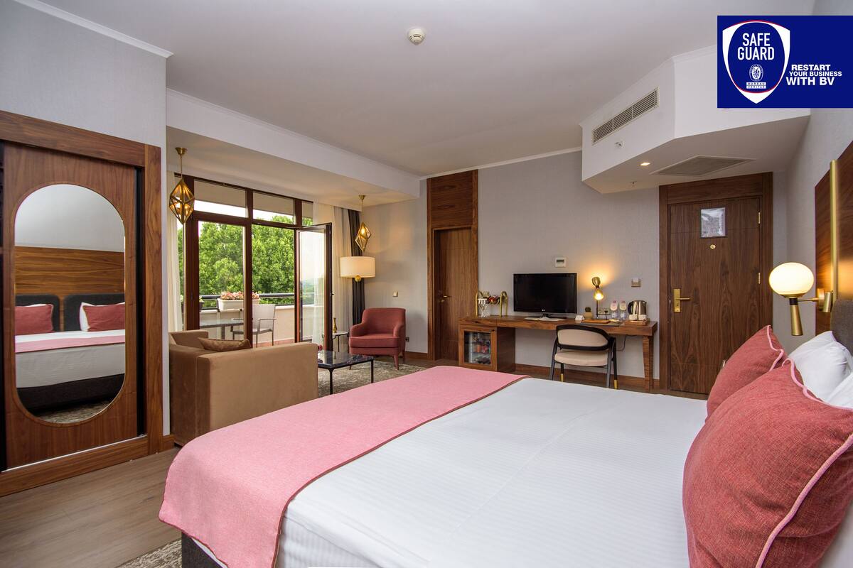 A hotel room with a large bed, a desk with a computer, a chair, a TV, and a window with a view of trees.