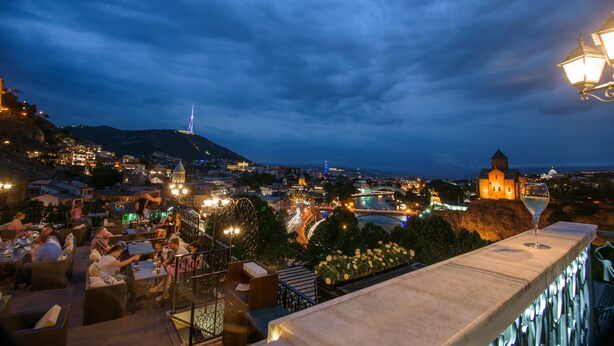 Eine Terrasse mit Sitzgelegenheiten im Freien mit Blick auf eine beleuchtete Stadtlandschaft bei Nacht, einschließlich erhellter Gebäude und eines entfernten Turms.