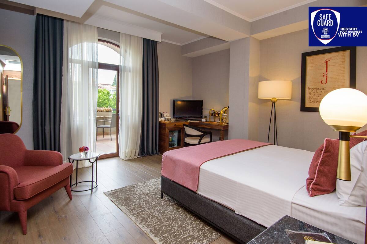A hotel room with a bed, a desk with a computer, a chair, a sofa, and a small table with a lamp.
