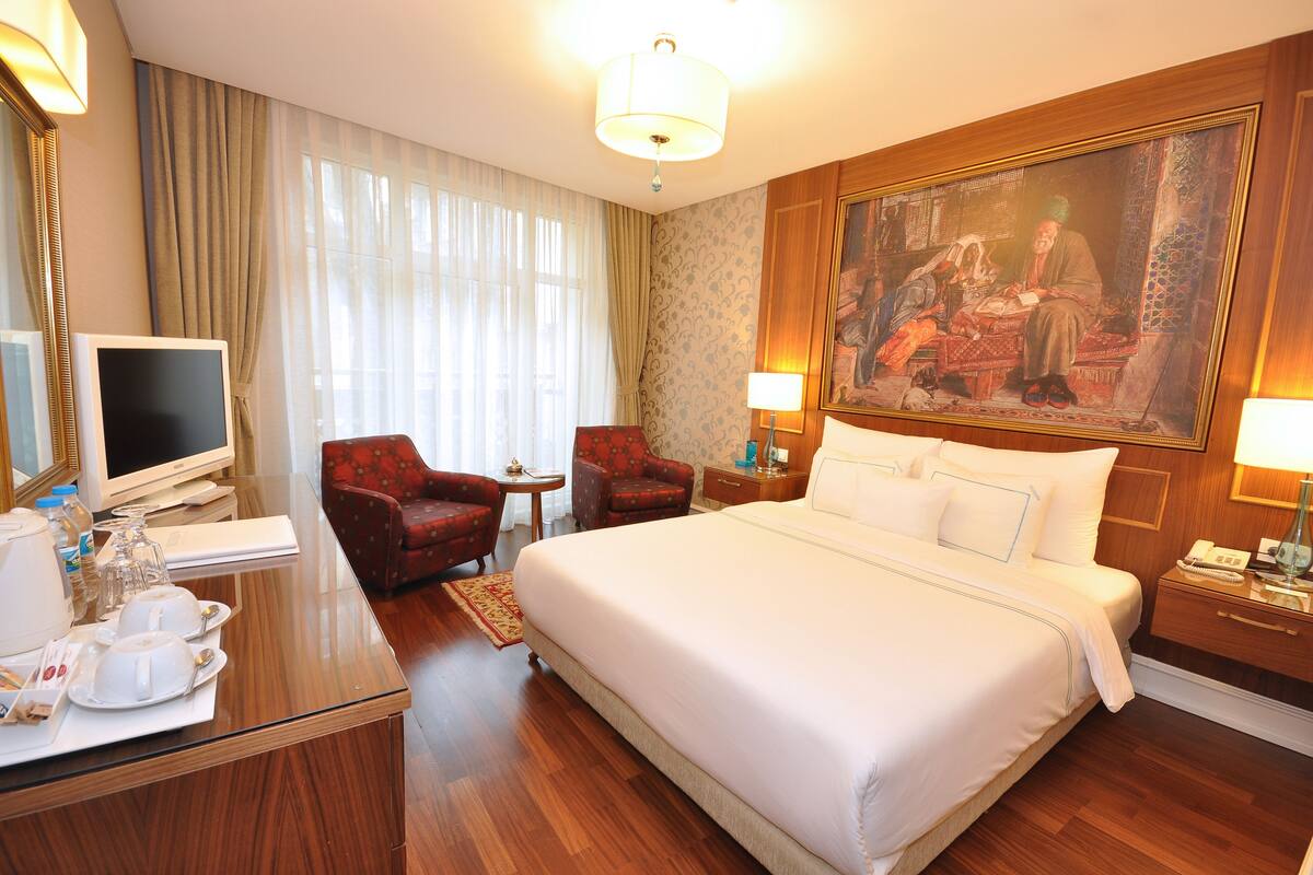 A hotel room with a large bed, a desk with a computer, two armchairs, a painting on the wall, and a nightstand with a lamp.