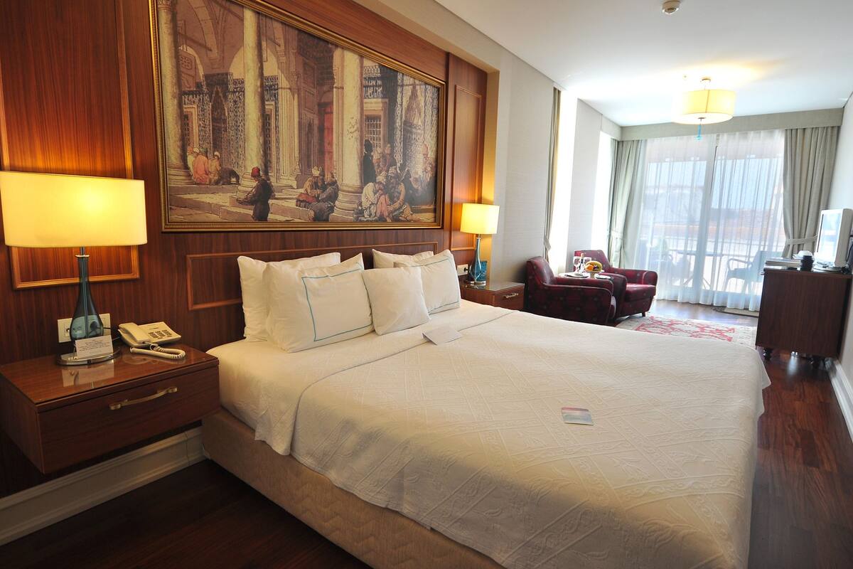 A hotel room with a large bed, two bedside tables with lamps, a desk with a chair, a large painting on the wall, and a view of the outdoors through the window.