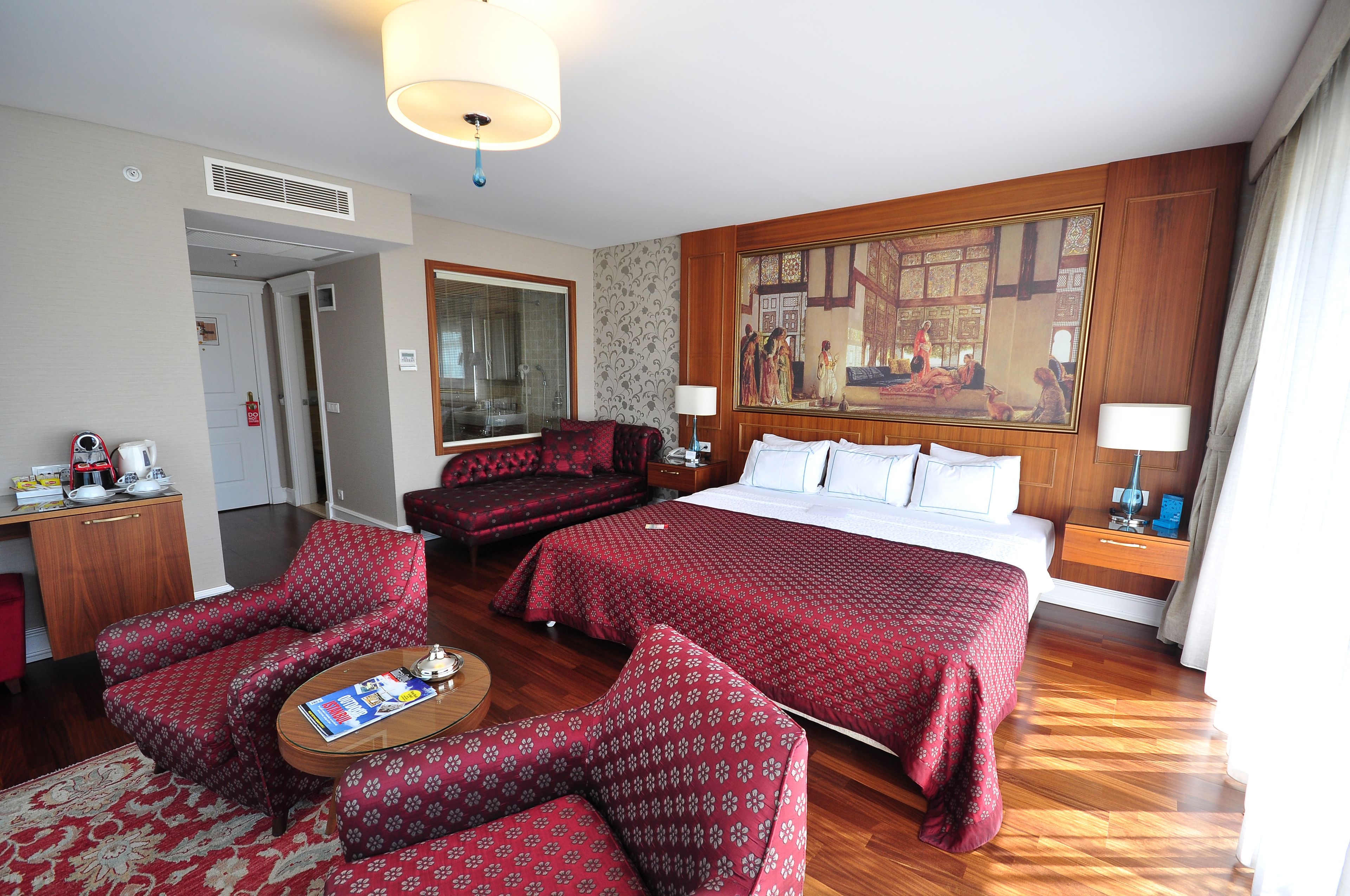 A hotel room with a large bed, two red patterned armchairs, a wooden desk with a coffee maker, and a large mirror.
