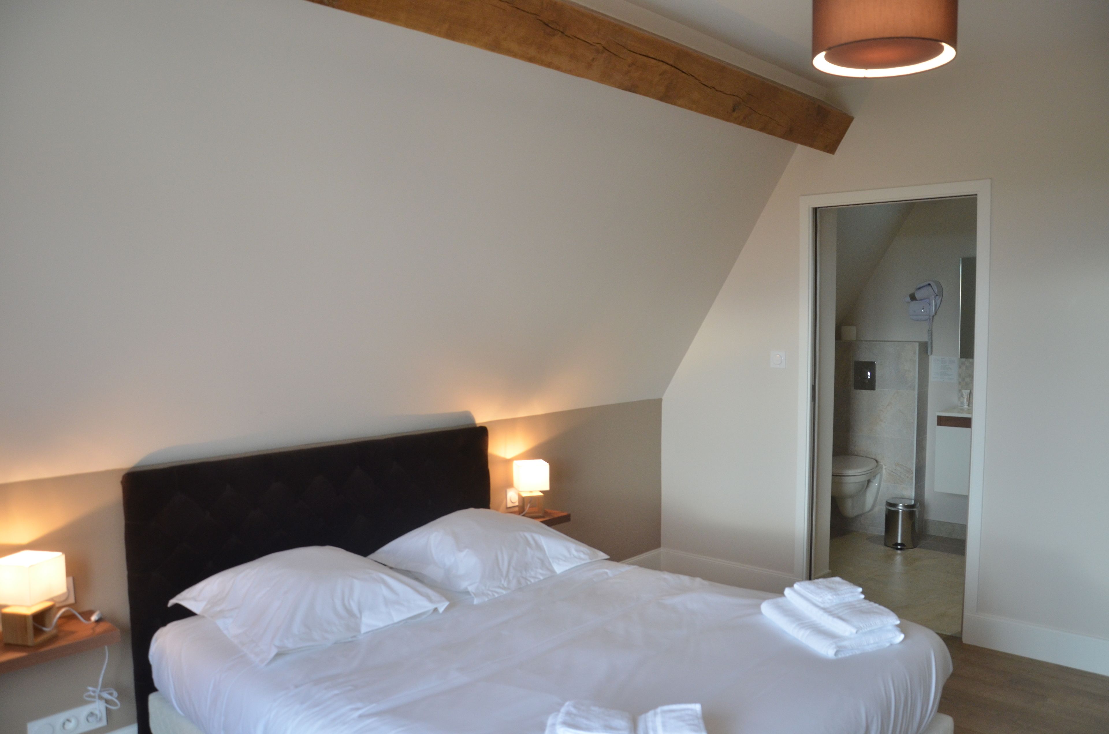 Een netjes opgemaakt bed met witte lakens en handdoeken, een houten hoofdbord, bedlampjes en uitzicht op de badkamer door een geopende deur.