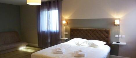 Quarto de hotel com uma cama grande, duas luminárias de cabeceira, um painel de cabeceira, uma janela com cortinas e um sofá.