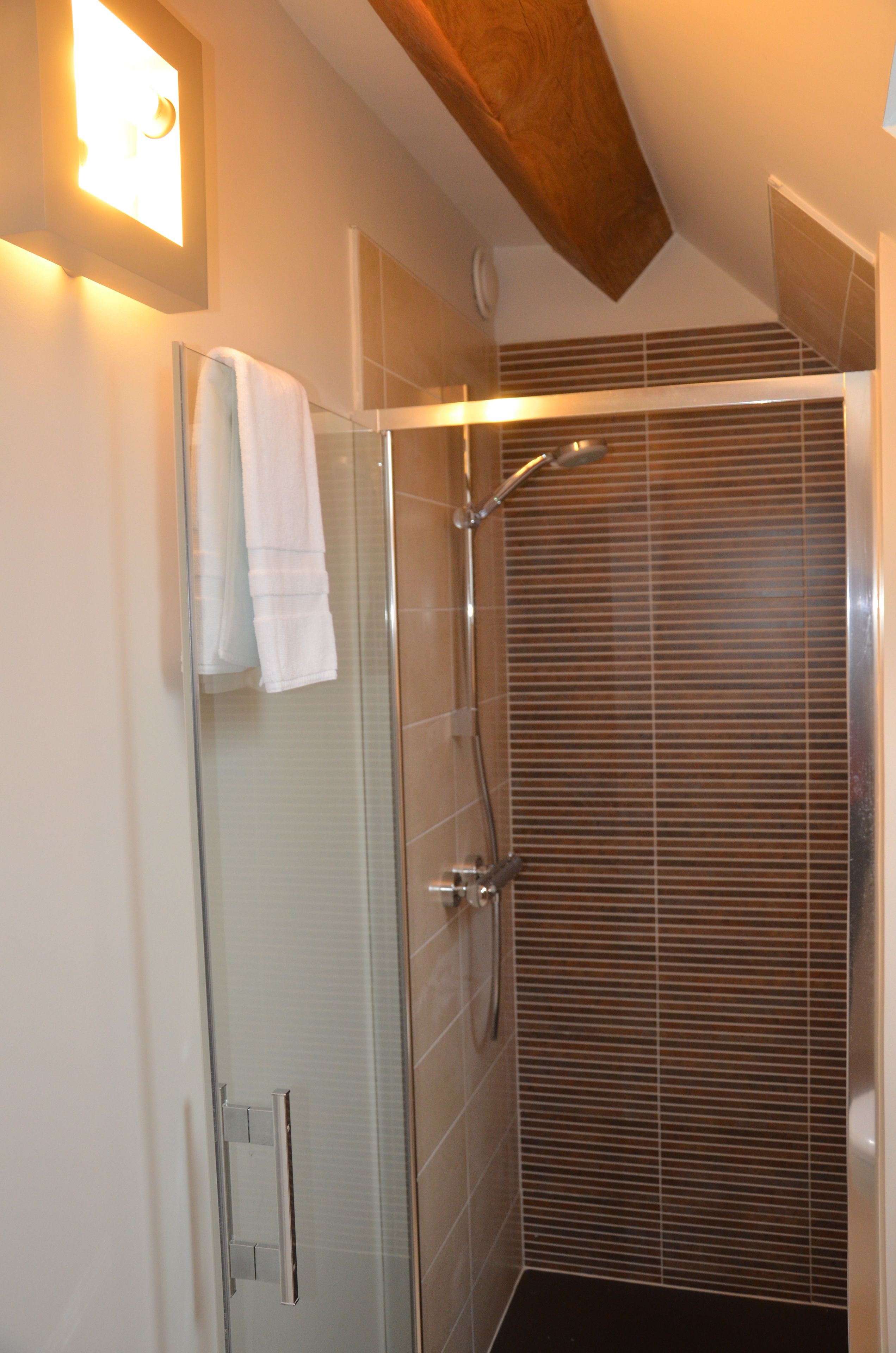 Eine moderne Dusche mit Glastür, ein weißes Handtuch am Türhänger und ein Holzbalken an der Decke.