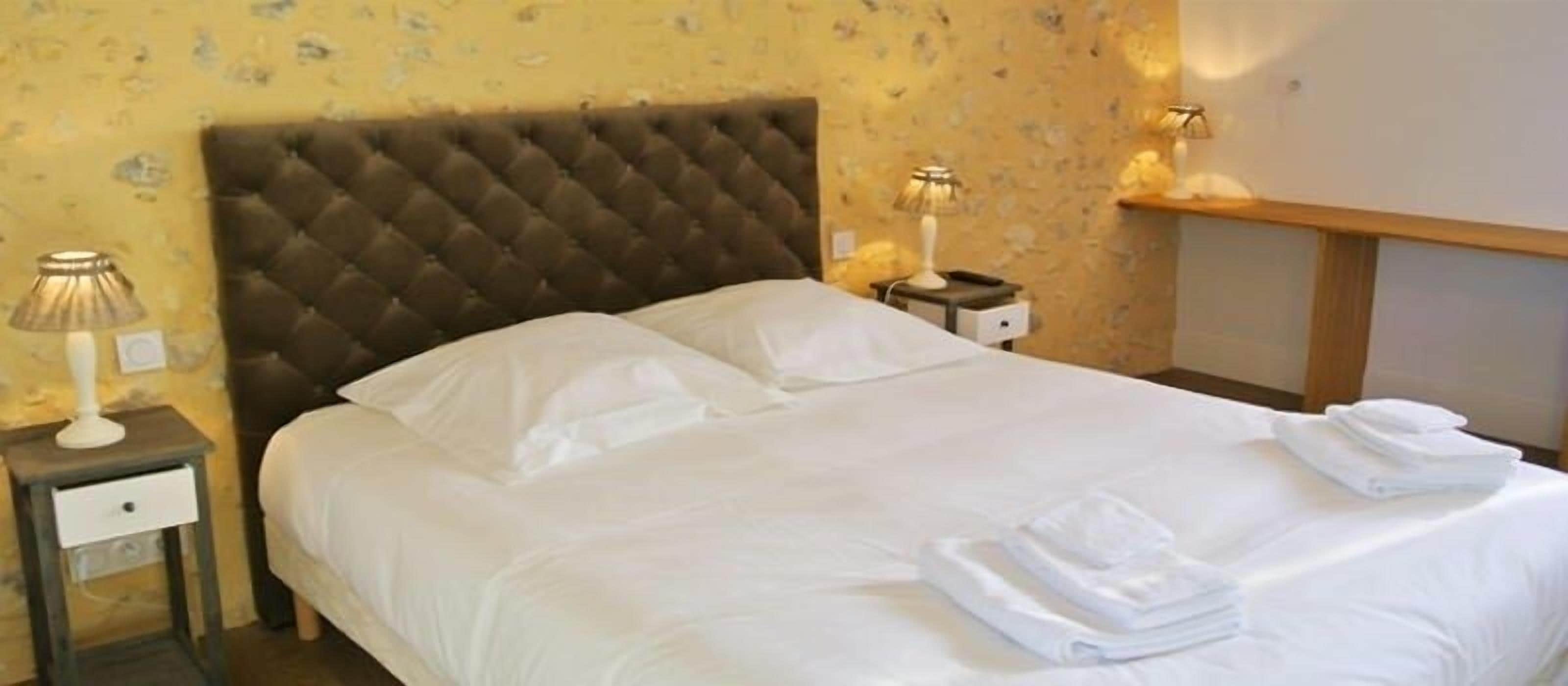 A neatly made bed with white linens and towels, a tufted headboard, a bedside lamp, and a small nightstand with a drawer.