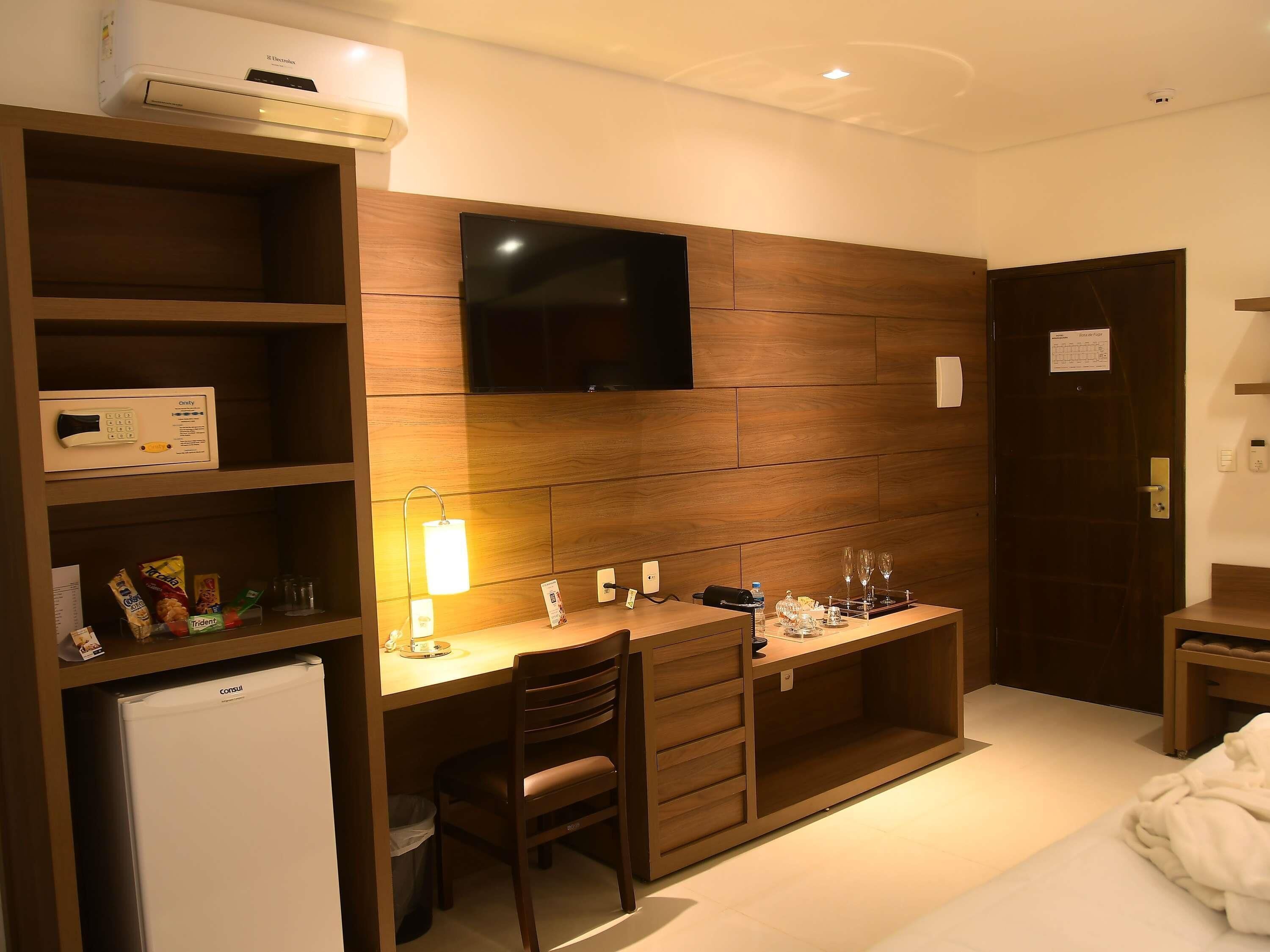 Quarto de hotel compacto com uma escrivaninha de madeira, televisão, minibar e uma cama.