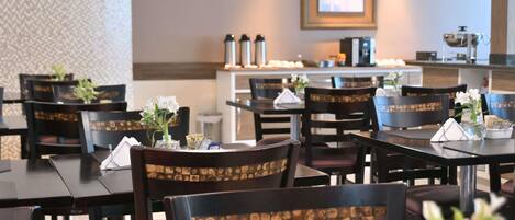 A restaurant with dark wooden tables and chairs, some with floral centerpieces. There are framed pictures on the wall and a coffee machine on the counter.