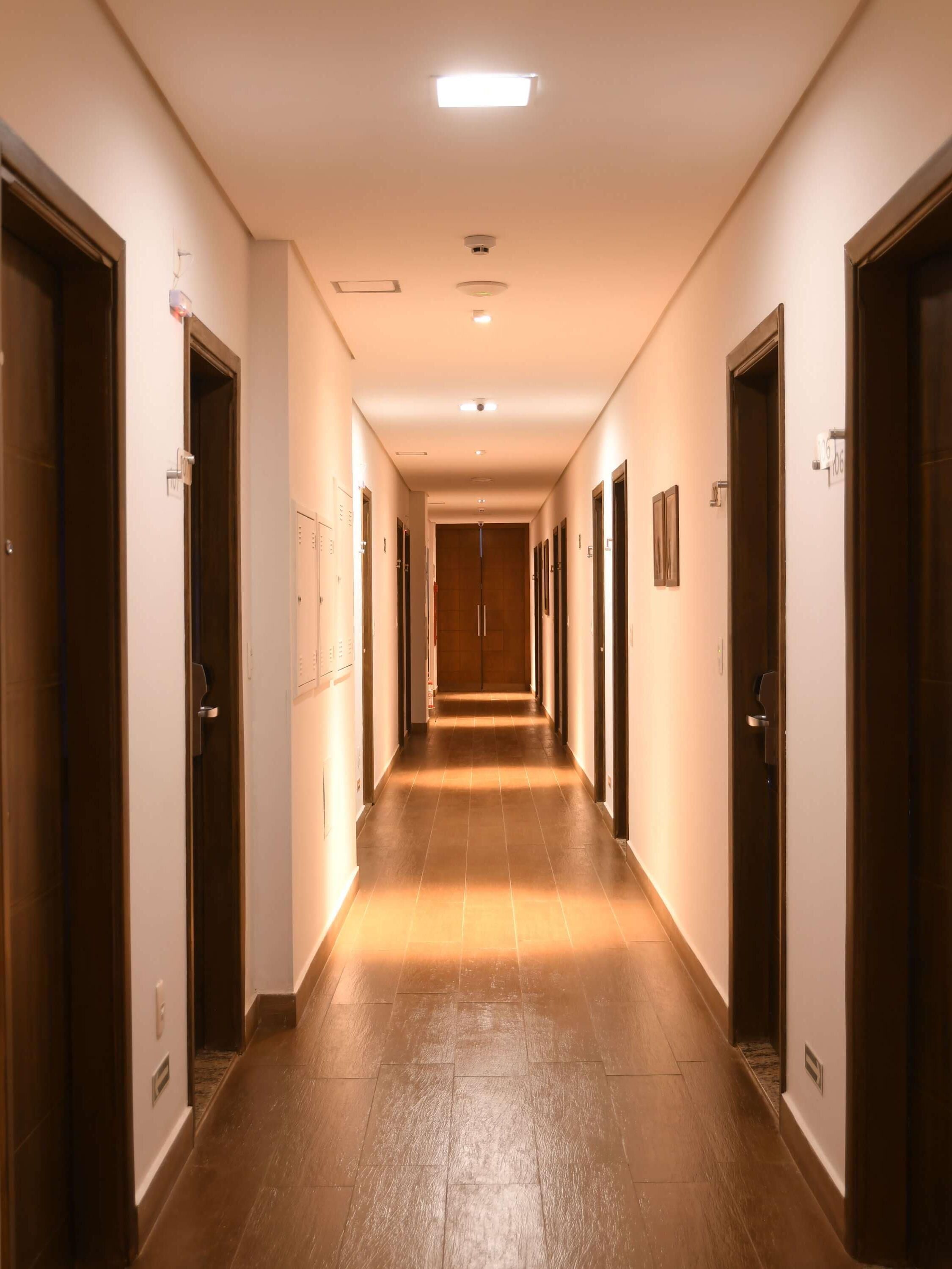Een lange, smalle hotelgangen met houten deuren aan beide zijden, een betegelde vloer en in het plafond weggewerkte verlichting.