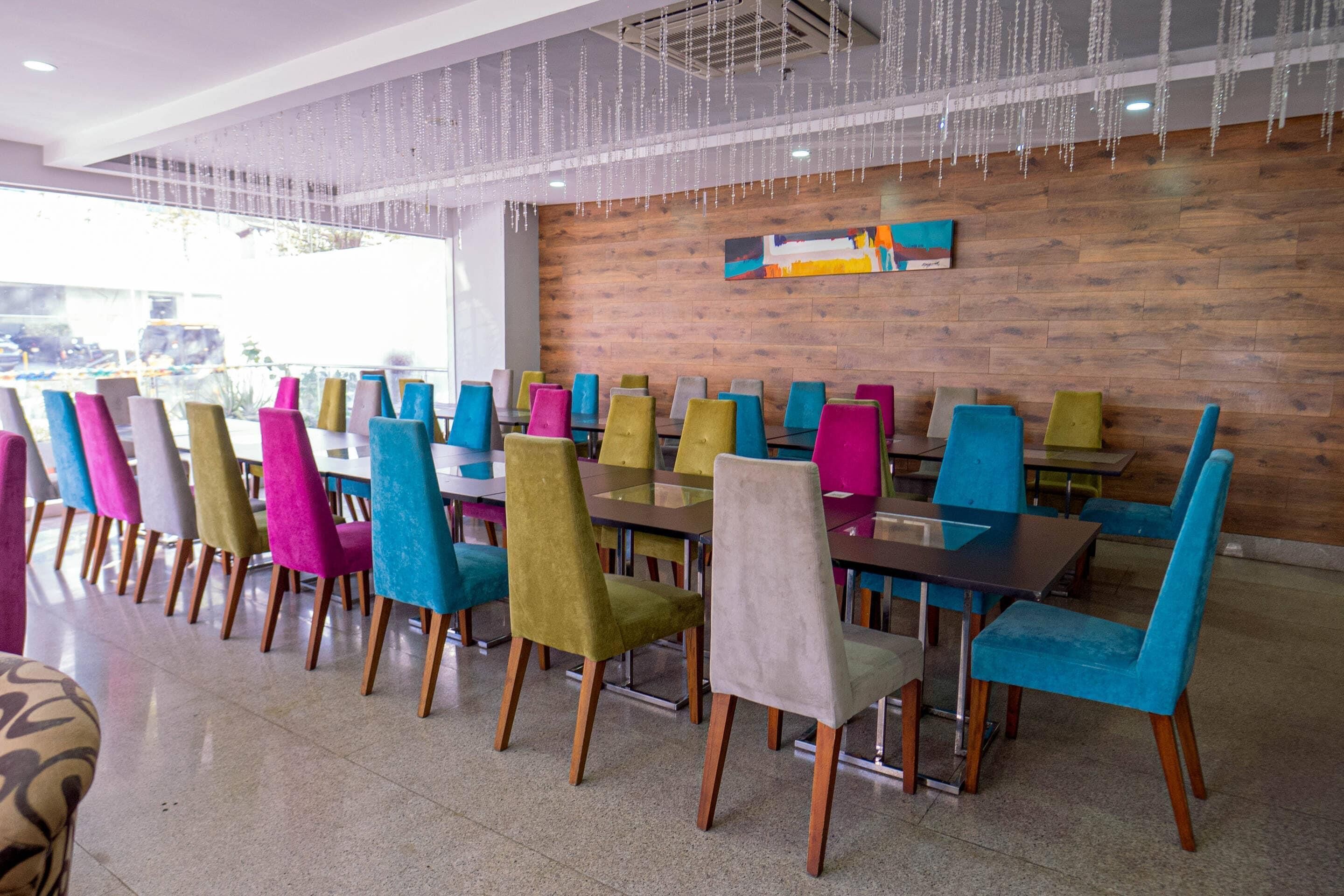 A dining area with multiple tables and chairs, a wooden wall with a colorful painting, and a ceiling with hanging lights.