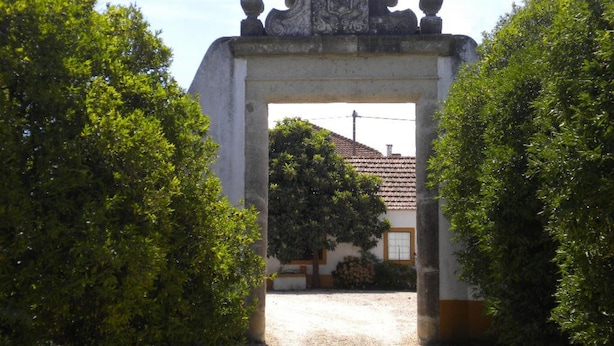 An entrance archway with a stone relief, flanked by lush green bushes, leading to a building with a tiled roof. - Quinta do Casalinho (Almeirim)