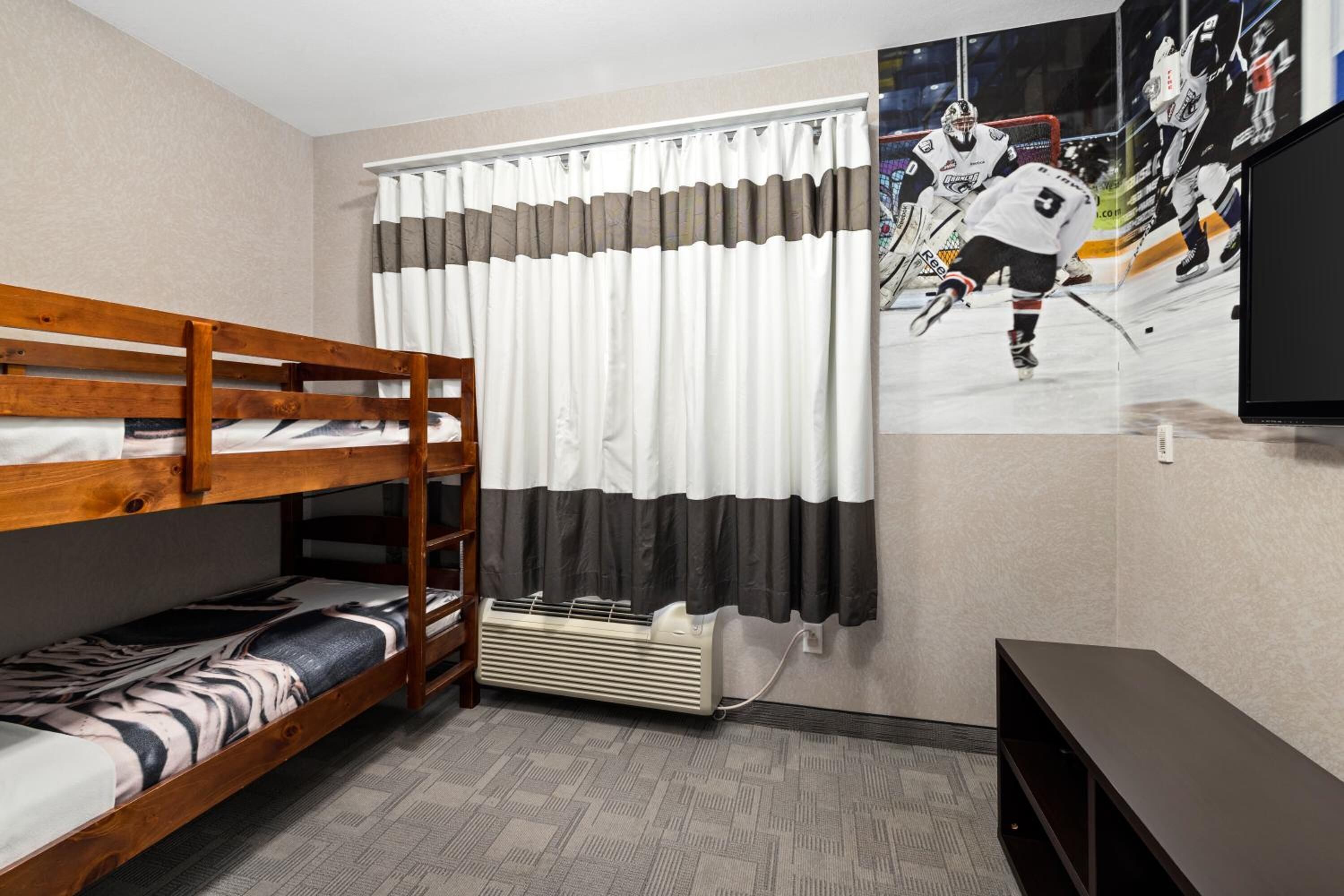 Un lit superposé avec une couvre-lit à motifs, un radiateur fixé au mur, une fenêtre avec des rideaux et une affiche sur le thème du hockey.