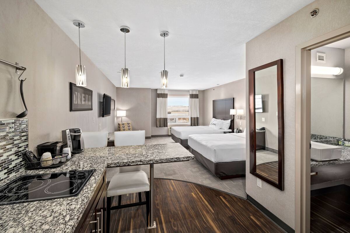 A hotel room with a kitchen area, a dining table, two beds, a large mirror, and a view of the outdoors.