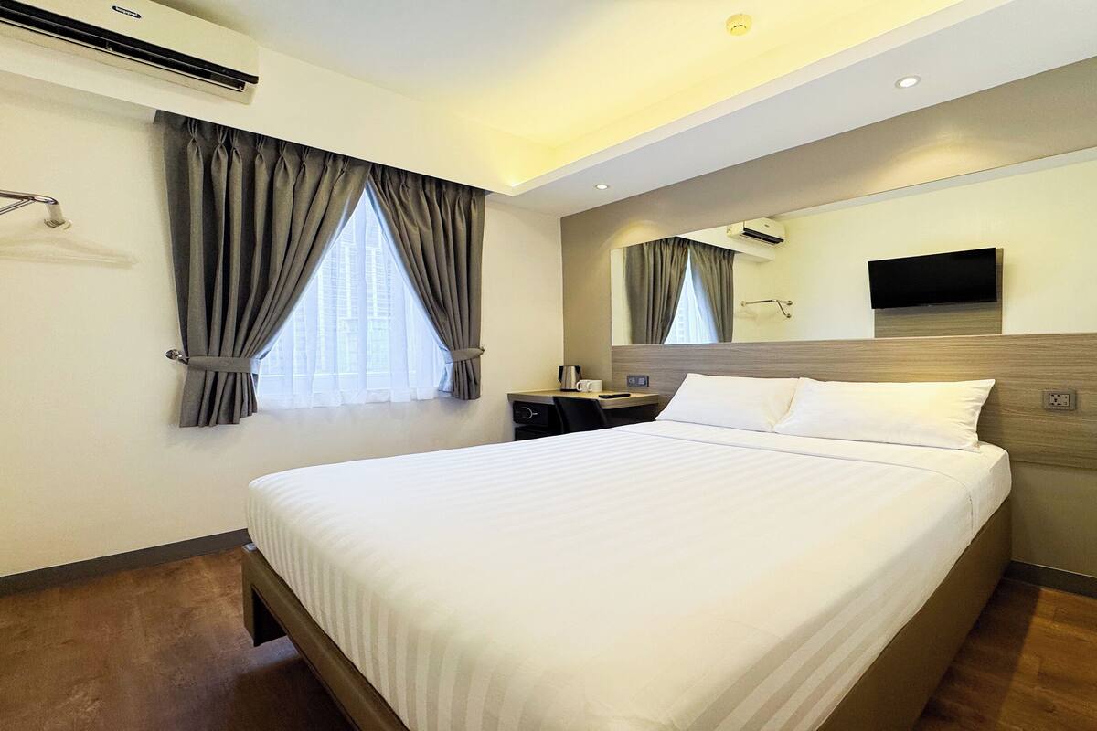 A hotel room with a large bed, a bedside table with a remote control, a flat-screen TV mounted on the wall, and an air conditioning unit on the wall.