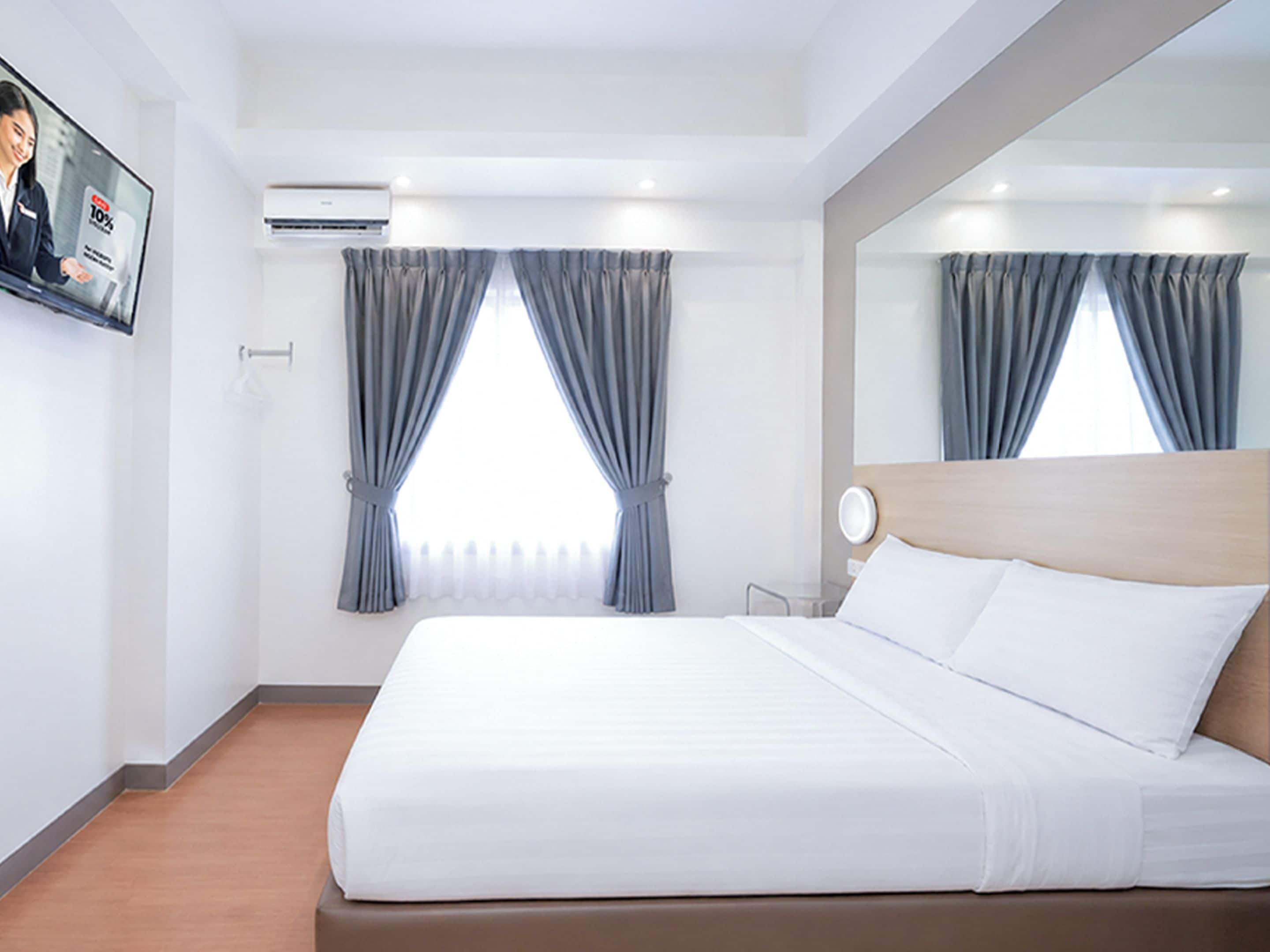 Habitación de hotel con cama, televisor montado en la pared, aire acondicionado y cortinas en las ventanas.