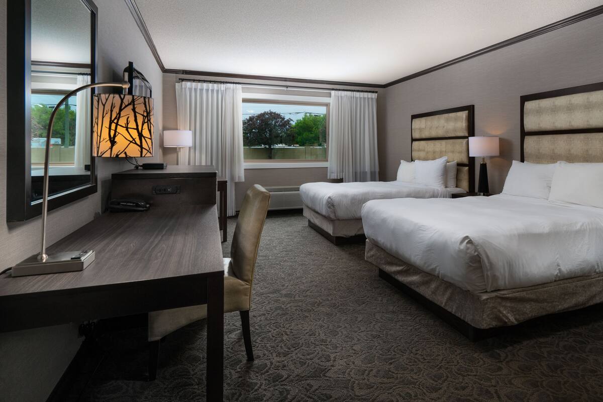 A hotel room with two beds, a desk, a chair, a lamp, and a window with curtains.